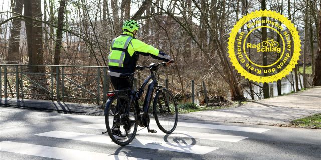 Ein Polizeibeamter in Fahrradkleidung schiebt ein Fahrrad über einen Zebrastreifen