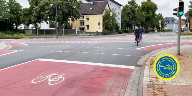 Straße in Gütersloh. Auf der Fahrbahn vor der Ampel befindet sich eine rote Aufstellfläche mit weißem Fahrradpiktogramm. In der Ferne sieht man zwei Radfahrende