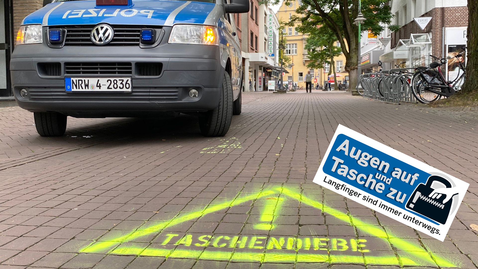 Blick in eine Fußgängerzone. Auf den Boden ist mit neonfarbiger Sprühkreide ein Warnzeichen mit der Aufschrift Taschendiebe gesprüht. Links im Bild steht ein Streifenwagen.