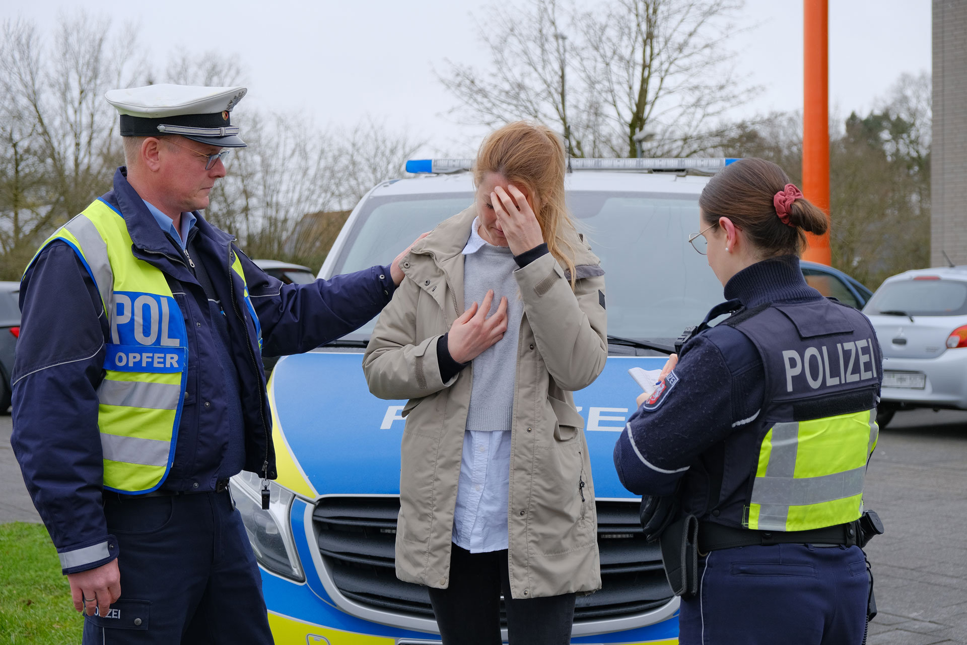 Eine Frau steht vor einem Streifenwagen, hält die Hand vors Gesicht und blickt nach unten. Ihre andere Hand liegt auf der Brust. Ein Polizist steht neben ihr und hat ihr die Hand auf die Schulter gelegt. Eine Polizistin steht vor ihr und macht sich Notizen