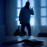 Ein dunkler Raum. Im Hintergrund sieht man einen Einbrecher mit Taschenlampe. Im Vordergrund liegt eine Geldbörse und ein Handy auf einem Tisch