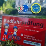 Schild mit der Aufschrift Radfahrprüfung. Im Hintergrund unscharf ein Junge auf einem Fahrrad beim Linksabbiegen. Er trägt Fahrradhelm und eine Weste in Neongelb mit der aufgedruckten Zahl 6