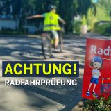 Schild mit der Aufschrift Radfahrprüfung. Im Hintergrund unscharf ein Junge auf einem Fahrrad beim Linksabbiegen. Er trägt Fahrradhelm und eine Weste in Neongelb mit der aufgedruckten Zahl 6. Headline: ACHTUNG! Radfahrprüfung