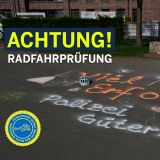 Ein Schulhof. Auf dem Boden ist mit Sprühkreide aufgesprüht "Viel Erfolg. Polizei Gütersloh" Neben dem Schriftzug liegt ein Fahrradhelm. Text "Achtung! Radfahrprüfung"