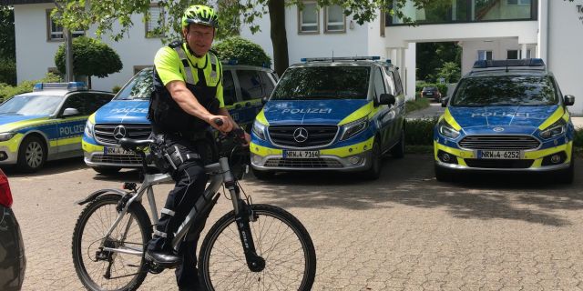 Polizeioberkommissar Markus Meyer zu Erpen in Fahrraduniform auf dem Fahrrad. Im Hintergrund drei Streifenwagen