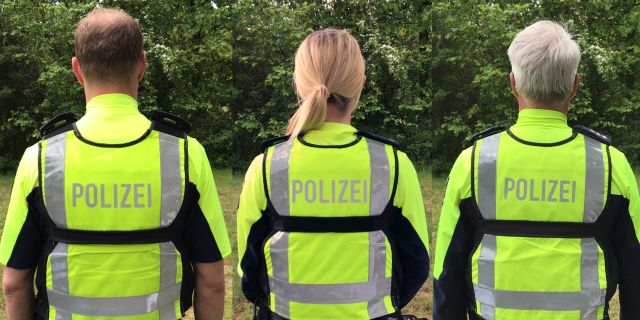 Rückenansicht von zwei Polizeibeamten und einer Polizeibeamtin. Alle drei tragen eine neonfarbene Polizei-Weste