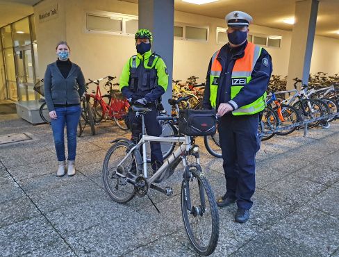 Zwei Polizeibeamte stehen neben einem Fahrrad. Sie tragen neonfarbene Bekleidung und Mund-Nase-Schutz. Neben Ihnen steht eine Frau, sie trägt ebenfalls einen Mund-Nase-Schutz. Kulisse sind die Fahrradständer des Kreisgymnasiums Halle.